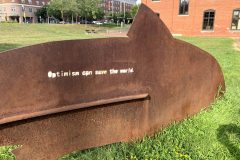 Metal fish art that says "Optimism can save the world"